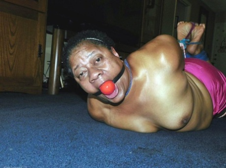 Overweight black woman Trixie struggles against a ball gag while hogtied